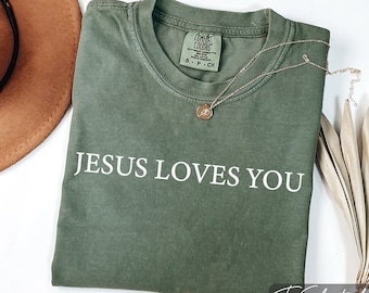 May include: Olive green t-shirt with the text "JESUS LOVES YOU" in white. A gold necklace with a pendant rests on the shirt. A brown hat and dried plant are in the background. The shirt is labeled "Comfort Colors".