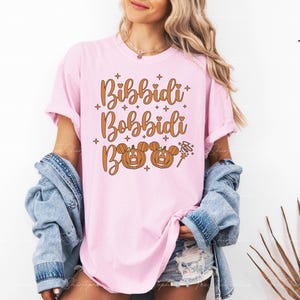 May include: A light pink t-shirt with the words "Bibbidi Bobbidi Boo" in a brown, whimsical font. The design includes two pumpkin Mickey Mouse heads and a magic wand. The shirt is part of a Halloween or Disney-themed collection.