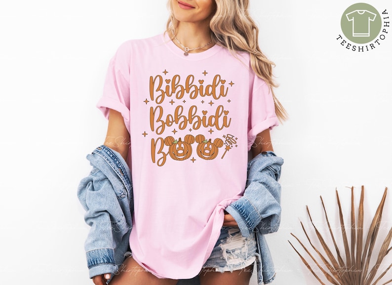 May include: A light pink t-shirt with the words "Bibbidi Bobbidi Boo" in a brown, whimsical font. The design includes two pumpkin Mickey Mouse heads and a magic wand. The shirt is part of a Halloween or Disney-themed collection.