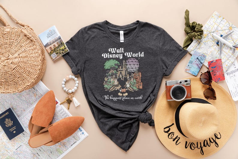 May include: Dark gray t-shirt with a Walt Disney World graphic and the text "The happiest place on earth." The image includes a straw hat, passport, camera, and other travel accessories.