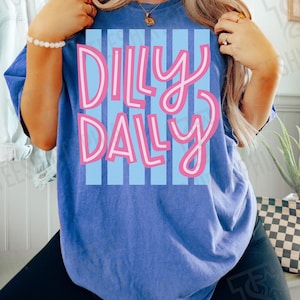 K&ouml;nnte beinhalten: Blaues T-Shirt mit dem Schriftzug "Dilly Dally" in Rosa, wei&szlig; umrandet, auf hellblau gestreiftem Hintergrund. Das T-Shirt hat eine einfarbige blaue Farbe.