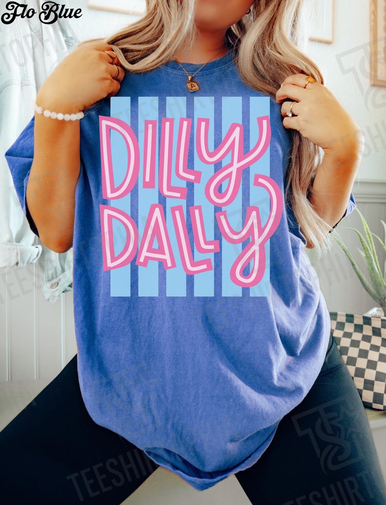 K&ouml;nnte beinhalten: Blaues T-Shirt mit dem Schriftzug "Dilly Dally" in Rosa, wei&szlig; umrandet, auf hellblau gestreiftem Hintergrund. Das T-Shirt hat eine einfarbige blaue Farbe.