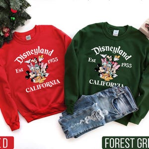 K&ouml;nnte beinhalten: Rote und waldgr&uuml;ne Sweatshirts mit dem Disneyland-Logo und Disney-Figuren. Die Sweatshirts tragen den wei&szlig;en Schriftzug "Disneyland Est 1955 California". Eine Jeans ist neben dem gr&uuml;nen Sweatshirt.
