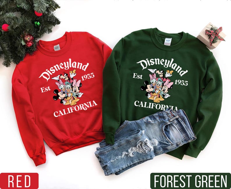K&ouml;nnte beinhalten: Rote und waldgr&uuml;ne Sweatshirts mit dem Disneyland-Logo und Disney-Figuren. Die Sweatshirts tragen den wei&szlig;en Schriftzug "Disneyland Est 1955 California". Eine Jeans ist neben dem gr&uuml;nen Sweatshirt.
