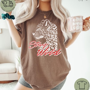 May include: Espresso-colored t-shirt featuring a white leopard graphic and the words "Stay Wild" in red script. The shirt has a relaxed fit and is made of a soft material. The shirt is part of the Teeshtophia collection.