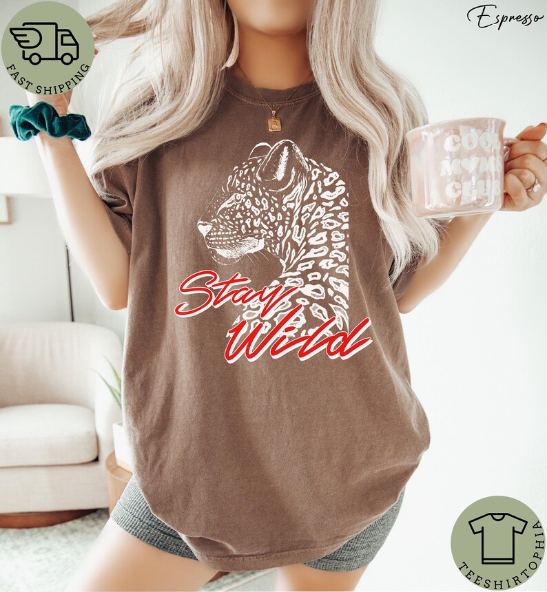 May include: Espresso-colored t-shirt featuring a white leopard graphic and the words "Stay Wild" in red script. The shirt has a relaxed fit and is made of a soft material. The shirt is part of the Teeshtophia collection.