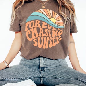 May include: Brown t-shirt with the text "FOREVER CHASING SUNSETS" in orange, with a sunset graphic in a circle. The graphic includes a blue wave, orange and yellow sun, and orange rays. The model is wearing blue jeans.