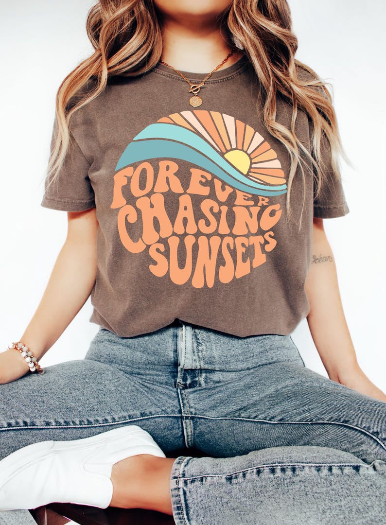 May include: Brown t-shirt with the text "FOREVER CHASING SUNSETS" in orange, with a sunset graphic in a circle. The graphic includes a blue wave, orange and yellow sun, and orange rays. The model is wearing blue jeans.