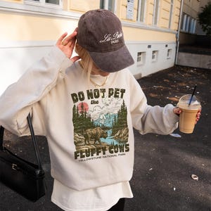 Puede incluir: Sudadera crema con el texto "DO NOT PET the FLUFFY COWS" y un gr&aacute;fico de un bisonte en un paisaje. Se combina con una gorra de b&eacute;isbol marr&oacute;n que dice "La Paris" en escritura blanca. El dise&ntilde;o est&aacute; inspirado en Yellowstone.
