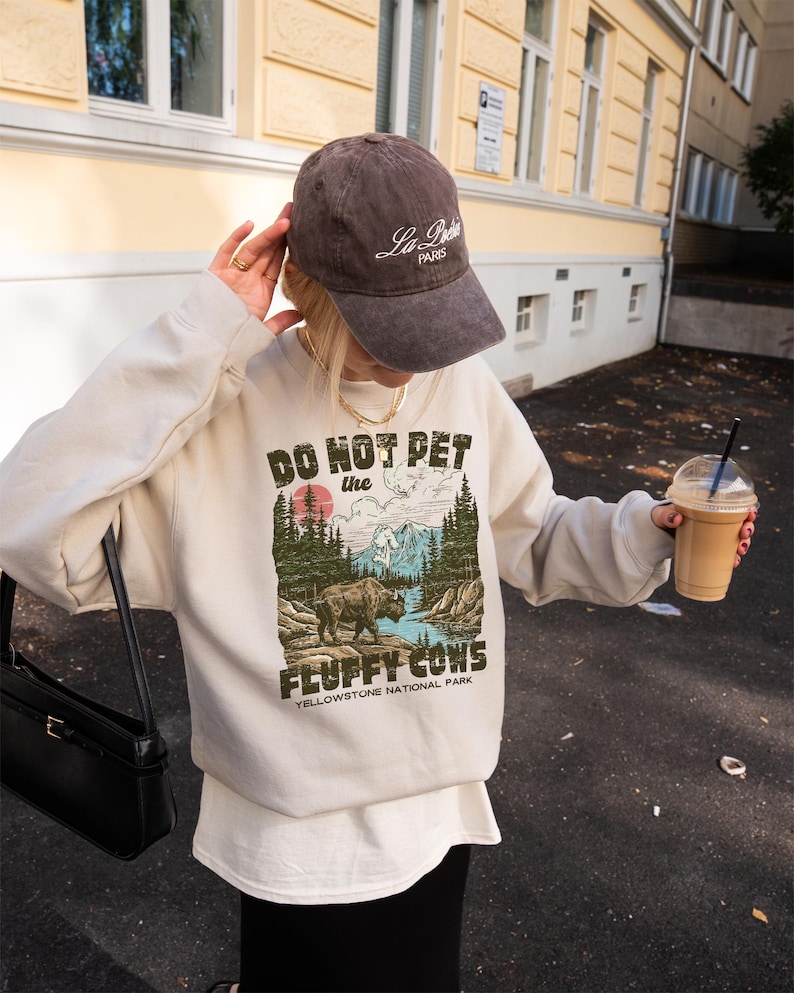 Puede incluir: Sudadera crema con el texto "DO NOT PET the FLUFFY COWS" y un gr&aacute;fico de un bisonte en un paisaje. Se combina con una gorra de b&eacute;isbol marr&oacute;n que dice "La Paris" en escritura blanca. El dise&ntilde;o est&aacute; inspirado en Yellowstone.