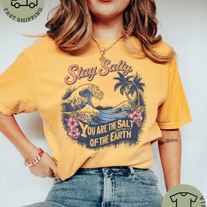 May include: Mustard-colored t-shirt with a graphic featuring the text "Stay Salty" above a wave, palm trees, and the text "You are the salt of the earth". The shirt has a vintage, distressed look.