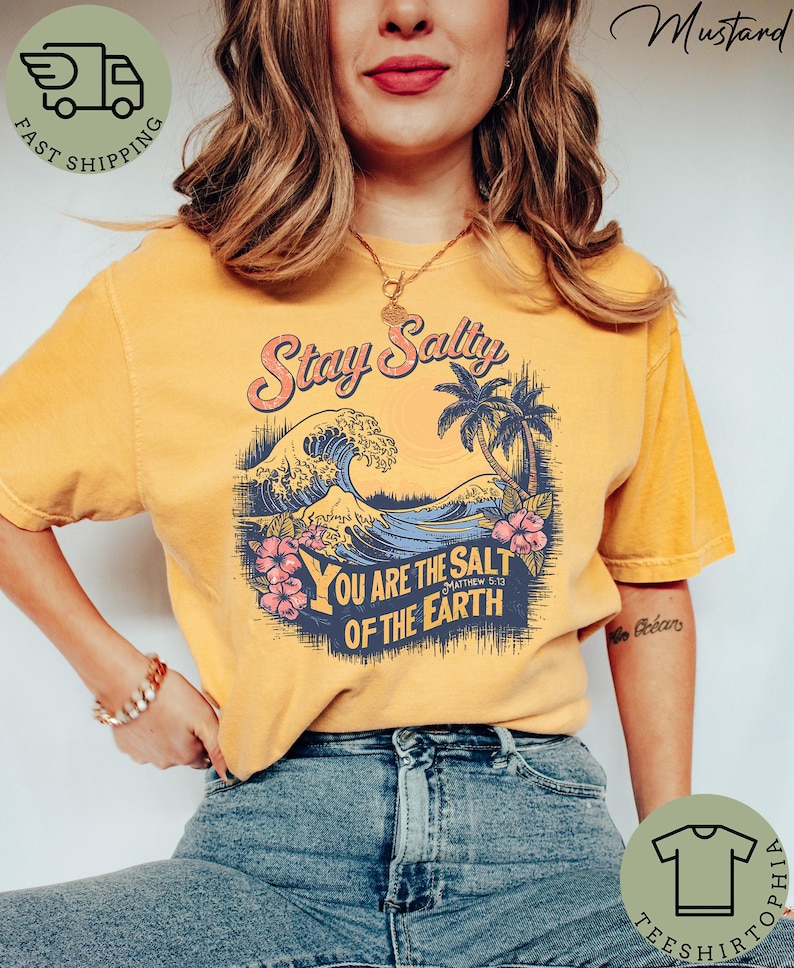 May include: Mustard-colored t-shirt with a graphic featuring the text "Stay Salty" above a wave, palm trees, and the text "You are the salt of the earth". The shirt has a vintage, distressed look.