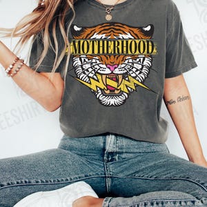 May include: Dark gray t-shirt featuring a tiger head graphic with the word "MOTHERHOOD" in a yellow banner. The tiger has orange and white stripes, pink nose, and a yellow lightning bolt. The shirt is paired with blue jeans and white sneakers.