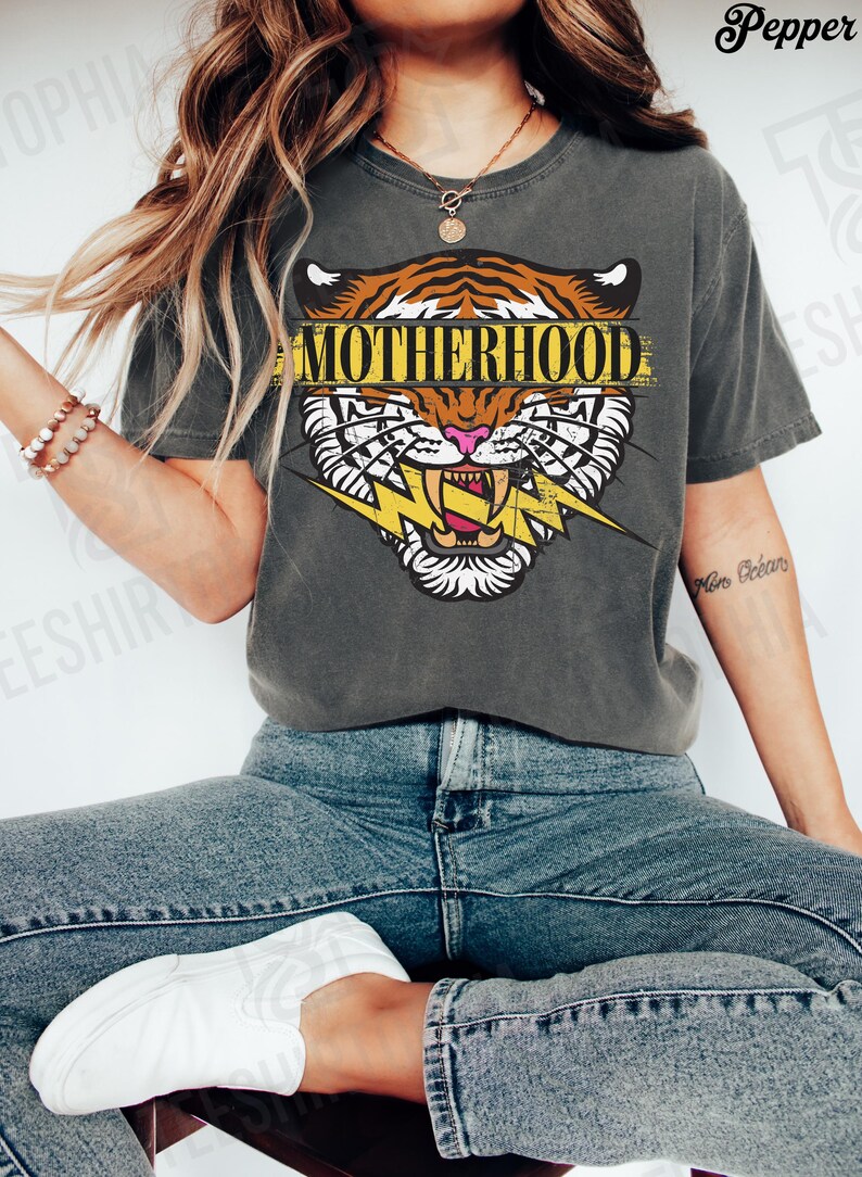 May include: Dark gray t-shirt featuring a tiger head graphic with the word "MOTHERHOOD" in a yellow banner. The tiger has orange and white stripes, pink nose, and a yellow lightning bolt. The shirt is paired with blue jeans and white sneakers.