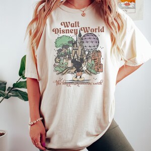 May include: Cream-colored t-shirt featuring a vintage-style graphic of Walt Disney World. The design includes a castle, Spaceship Earth, Mickey Mouse, and the text "Walt Disney World" and "the happiest place on earth."