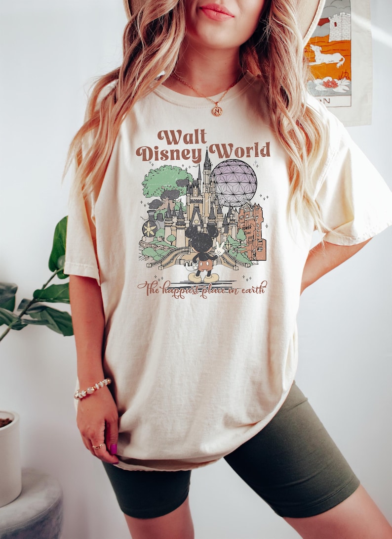 May include: Cream-colored t-shirt featuring a vintage-style graphic of Walt Disney World. The design includes a castle, Spaceship Earth, Mickey Mouse, and the text "Walt Disney World" and "the happiest place on earth."