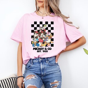 May include: Light pink t-shirt featuring a group of cartoon characters in front of a black and white checkered background. The text "Mickey & Co. EST. 1928" is printed below the characters. The shirt is worn by a person.