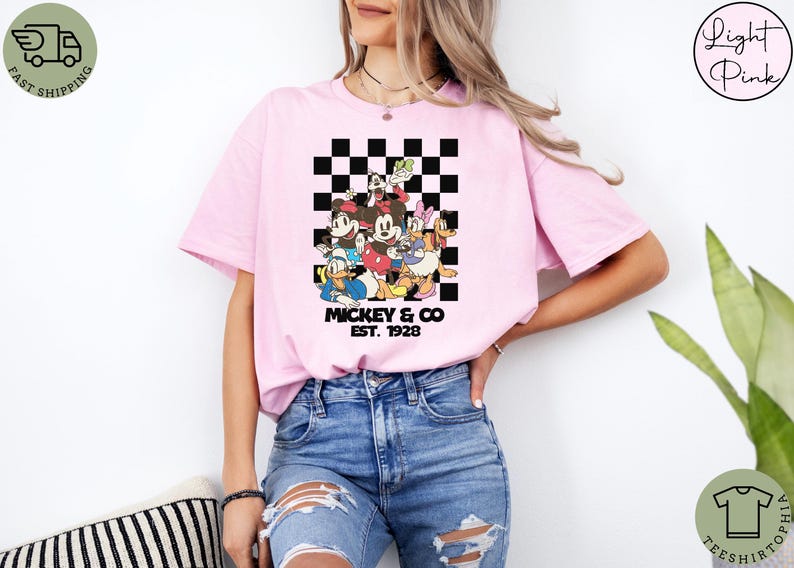 May include: Light pink t-shirt featuring a group of cartoon characters in front of a black and white checkered background. The text "Mickey & Co. EST. 1928" is printed below the characters. The shirt is worn by a person.
