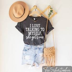 May include: Dark gray t-shirt with the text "I LOVE TALKING TO MYSELF. she gets me." paired with denim shorts, a tan hat, and a woven crossbody bag. The shirt is hanging on a wooden hanger.