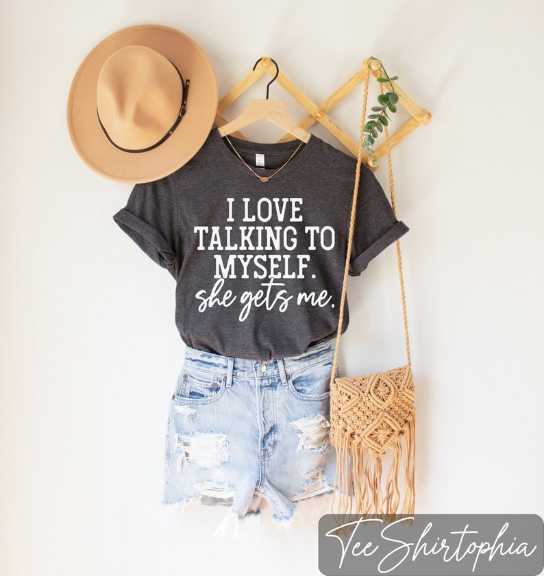 May include: Dark gray t-shirt with the text "I LOVE TALKING TO MYSELF. she gets me." paired with denim shorts, a tan hat, and a woven crossbody bag. The shirt is hanging on a wooden hanger.