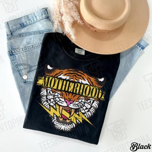 May include: Black t-shirt with a tiger graphic and the word "MOTHERHOOD" in yellow. The shirt is paired with light blue jeans and a tan hat. A gold necklace is visible.