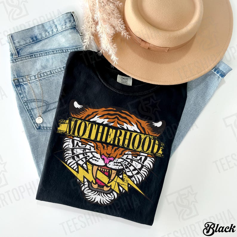 May include: Black t-shirt with a tiger graphic and the word "MOTHERHOOD" in yellow. The shirt is paired with light blue jeans and a tan hat. A gold necklace is visible.