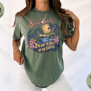 May include: Moss green t-shirt with a graphic design featuring a wave, palm trees, and the text "Stay Salty You Are The Salt Of The Earth". The design includes pink, blue, and yellow accents. The shirt is a casual wear item.