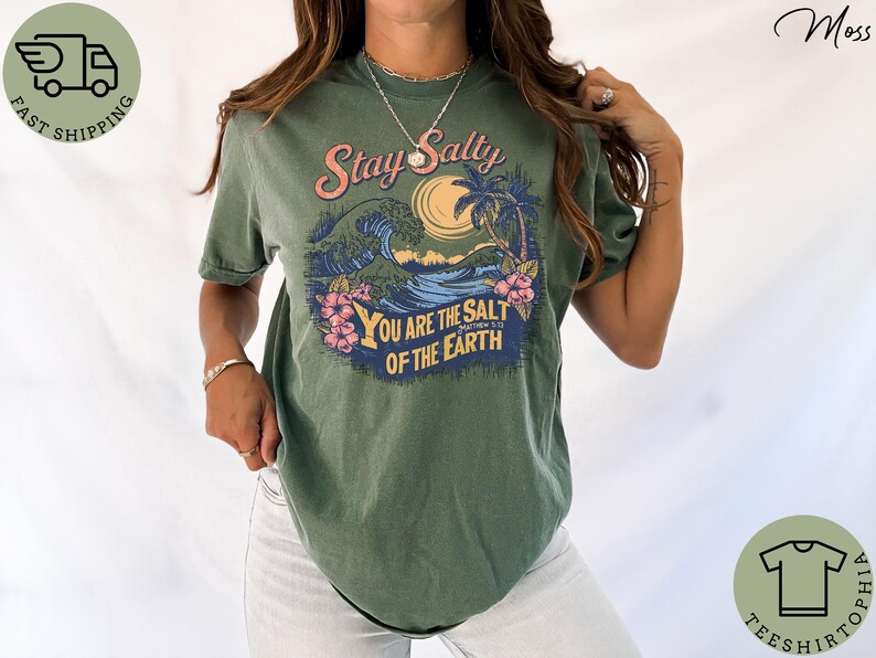 May include: Moss green t-shirt with a graphic design featuring a wave, palm trees, and the text "Stay Salty You Are The Salt Of The Earth". The design includes pink, blue, and yellow accents. The shirt is a casual wear item.