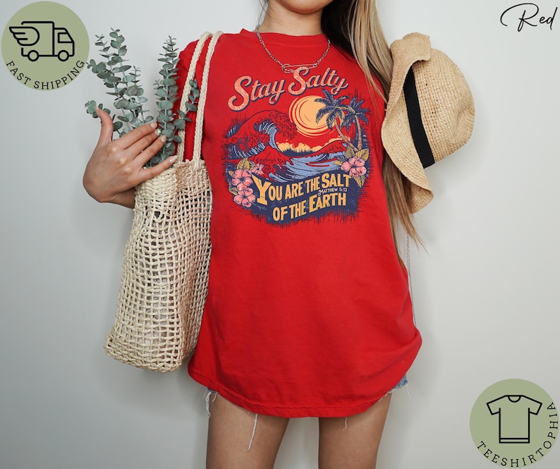 May include: Red t-shirt with a graphic design featuring a sunset, ocean waves, and palm trees. The text "Stay Salty" and "You are the salt of the earth" are included. A straw hat and a woven tote bag are also visible.