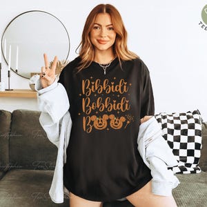 May include: Black t-shirt with the words "Bibbidi Bobbidi Boo" in orange script, along with Mickey Mouse jack-o'-lantern faces. The shirt is worn with a light-wash denim jacket. A peace sign is made with the fingers.