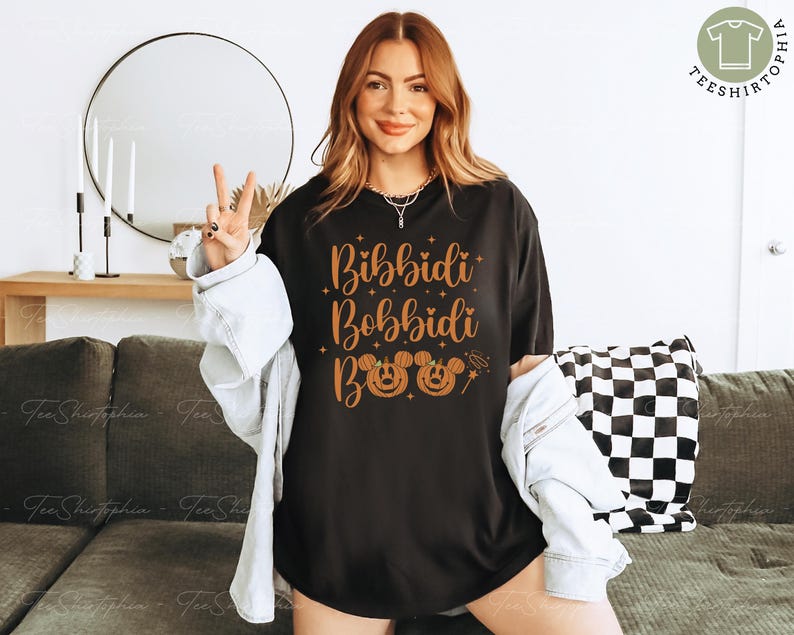 May include: Black t-shirt with the words "Bibbidi Bobbidi Boo" in orange script, along with Mickey Mouse jack-o'-lantern faces. The shirt is worn with a light-wash denim jacket. A peace sign is made with the fingers.