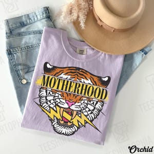 May include: A lilac-colored t-shirt with a tiger graphic and the word "MOTHERHOOD" in yellow. The tiger has orange and black stripes, a pink nose, and a yellow lightning bolt. The shirt is paired with blue jeans and a tan hat.