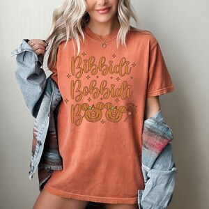 May include: Orange t-shirt with the text "Bibbidi Bobbidi Boo" in a whimsical font. The design includes pumpkin faces and stars, perfect for Halloween or a Disney-themed event.