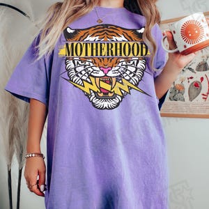 May include: A violet t-shirt with a tiger graphic and the word "MOTHERHOOD". The tiger design features orange and black stripes, a pink nose, and a yellow lightning bolt. The shirt is a relaxed fit.
