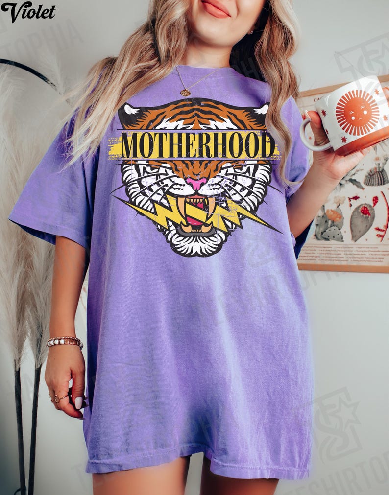 May include: A violet t-shirt with a tiger graphic and the word "MOTHERHOOD". The tiger design features orange and black stripes, a pink nose, and a yellow lightning bolt. The shirt is a relaxed fit.