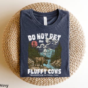 Puede incluir: Camiseta azul marino con un gr&aacute;fico que presenta un bisonte en un paisaje del Parque Nacional de Yellowstone. El texto dice "DO NOT PET the FLUFFY COWS". El dise&ntilde;o incluye un sol rojo, monta&ntilde;as y &aacute;rboles.