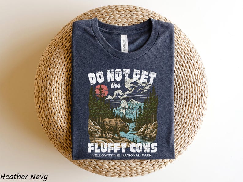 Puede incluir: Camiseta azul marino con un gr&aacute;fico que presenta un bisonte en un paisaje del Parque Nacional de Yellowstone. El texto dice "DO NOT PET the FLUFFY COWS". El dise&ntilde;o incluye un sol rojo, monta&ntilde;as y &aacute;rboles.