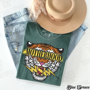 May include: A green t-shirt with a tiger graphic and the word "MOTHERHOOD" in yellow. The shirt is paired with light blue jeans and a tan hat. A gold necklace is visible.