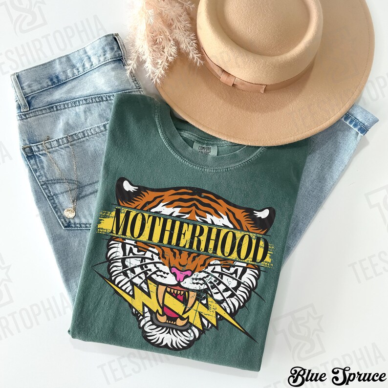 May include: A green t-shirt with a tiger graphic and the word "MOTHERHOOD" in yellow. The shirt is paired with light blue jeans and a tan hat. A gold necklace is visible.