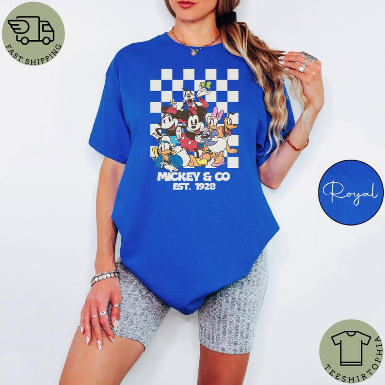 May include: Royal blue t-shirt featuring a graphic of Mickey & Co. characters with a checkered background. The text "Mickey & Co. Est. 1928" is printed below the characters. The shirt is a vibrant blue color.