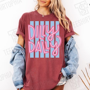 K&ouml;nnte beinhalten: Ein weinrotes T-Shirt mit dem Schriftzug "Dilly Dally" in rosa Schrift &uuml;ber hellblauen vertikalen Streifen. Das T-Shirt hat hochgekrempelte &Auml;rmel und wird mit einer Jeansjacke getragen. Es ist ein Freizeitkleidungsst&uuml;ck.