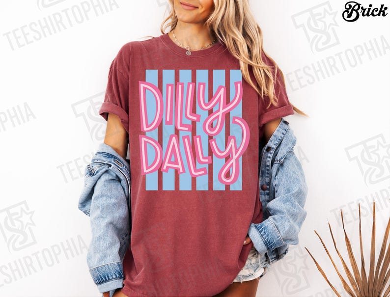 K&ouml;nnte beinhalten: Ein weinrotes T-Shirt mit dem Schriftzug "Dilly Dally" in rosa Schrift &uuml;ber hellblauen vertikalen Streifen. Das T-Shirt hat hochgekrempelte &Auml;rmel und wird mit einer Jeansjacke getragen. Es ist ein Freizeitkleidungsst&uuml;ck.