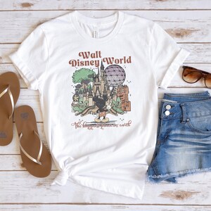 May include: White t-shirt with a graphic of Walt Disney World, featuring Mickey Mouse, a castle, and the text "The happiest place on earth." The shirt is paired with denim shorts, brown sandals, and sunglasses.