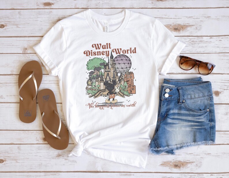 May include: White t-shirt with a graphic of Walt Disney World, featuring Mickey Mouse, a castle, and the text "The happiest place on earth." The shirt is paired with denim shorts, brown sandals, and sunglasses.
