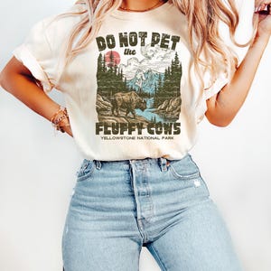 Puede incluir: Camiseta color arena con el texto "DO NOT PET the FLUFFY COWS" sobre un gr&aacute;fico de un bisonte en el Parque Nacional de Yellowstone. El gr&aacute;fico incluye un sol rojo, monta&ntilde;as y &aacute;rboles. La camiseta se combina con vaqueros azul claro.