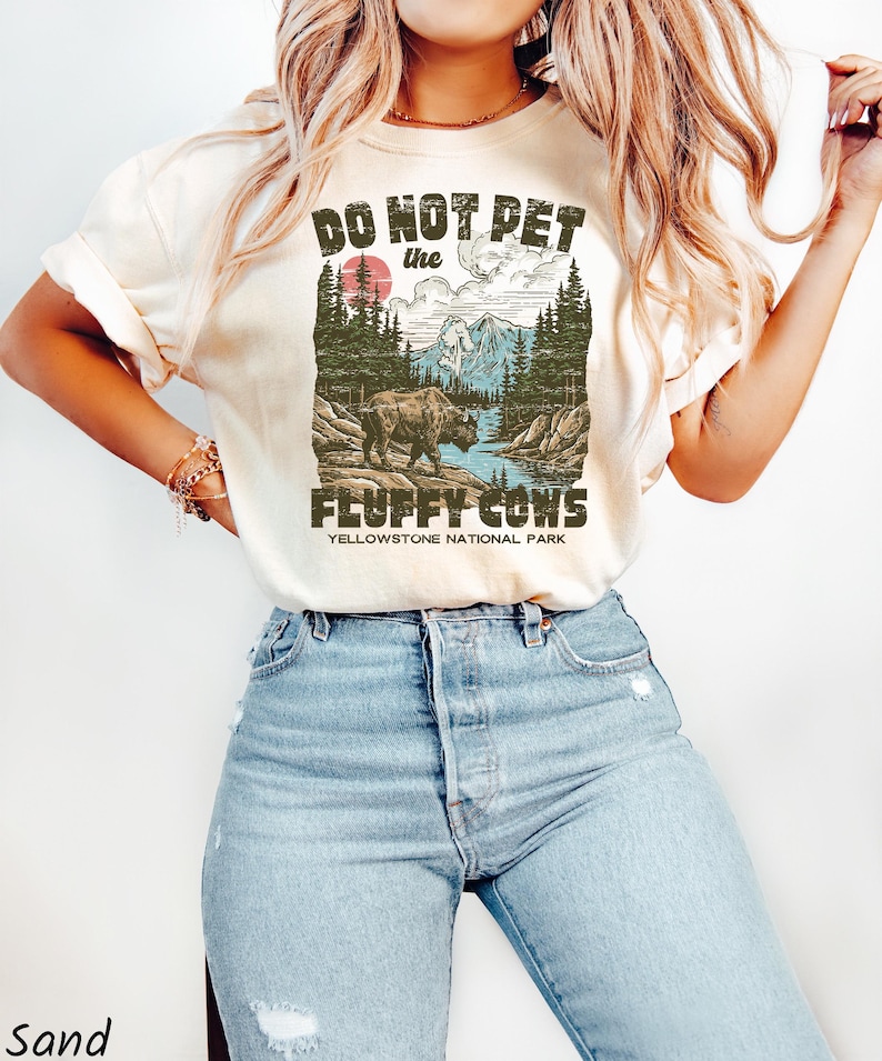 Puede incluir: Camiseta color arena con el texto "DO NOT PET the FLUFFY COWS" sobre un gr&aacute;fico de un bisonte en el Parque Nacional de Yellowstone. El gr&aacute;fico incluye un sol rojo, monta&ntilde;as y &aacute;rboles. La camiseta se combina con vaqueros azul claro.
