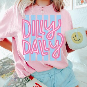 K&ouml;nnte beinhalten: Ein hellrosa T-Shirt mit den Worten "Dilly Dally" in rosa Schrift &uuml;ber hellblauen vertikalen Streifen. Eine wei&szlig;e Tasse mit einem gelben Smiley wird in der Hand gehalten. Die Person tr&auml;gt hellblaue Shorts.