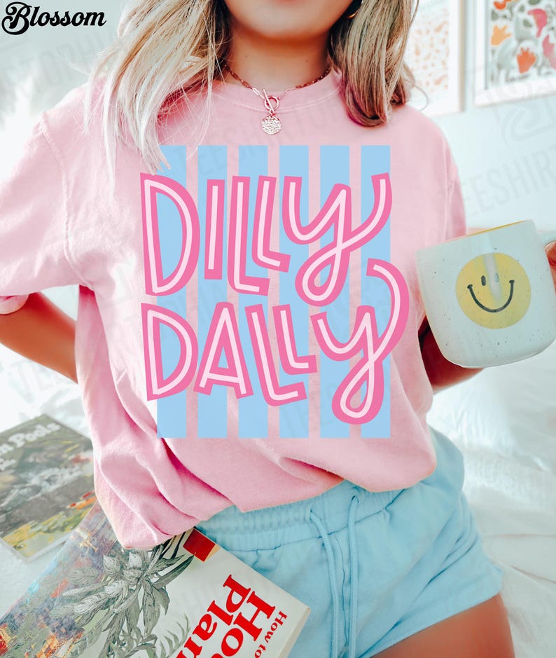 K&ouml;nnte beinhalten: Ein hellrosa T-Shirt mit den Worten "Dilly Dally" in rosa Schrift &uuml;ber hellblauen vertikalen Streifen. Eine wei&szlig;e Tasse mit einem gelben Smiley wird in der Hand gehalten. Die Person tr&auml;gt hellblaue Shorts.