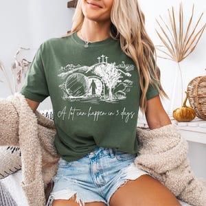 Op de afbeelding: Olijfgroen t-shirt met een witte afbeelding van een graf en de tekst "A lot can happen in 3 days." Het shirt is gemaakt van een zacht materiaal en is geschikt voor vrijetijdskleding. Het ontwerp heeft een religieus thema.