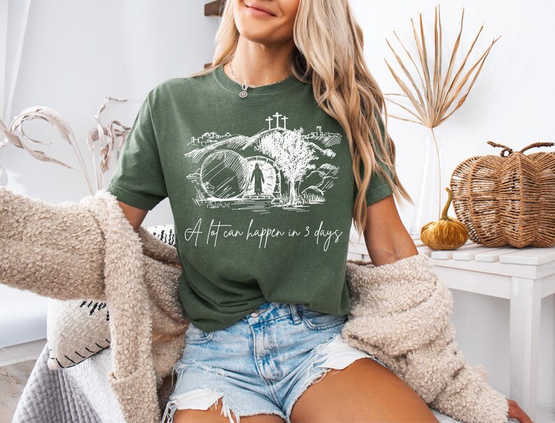 Op de afbeelding: Olijfgroen t-shirt met een witte afbeelding van een graf en de tekst "A lot can happen in 3 days." Het shirt is gemaakt van een zacht materiaal en is geschikt voor vrijetijdskleding. Het ontwerp heeft een religieus thema.